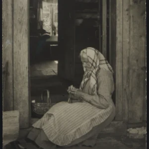 Photograph by Olaf Christoffersen of a rural woman knitting, framed by a doorway that offers a glimpse into the warmth and rhythm of her daily life.