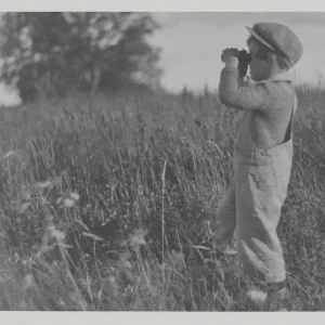 Photograph by Olaf Christoffersen of a young boy standing in a meadow, using binoculars to explore his surroundings, evoking curiosity and the spirit of childhood.