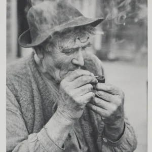 Photograph by Olaf Christoffersen of an elderly man lighting his pipe, highlighting his weathered face, rough hands, and the calm, contemplative atmosphere of the moment.