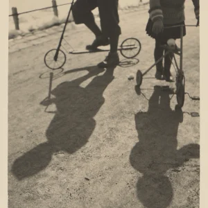 Photograph by Olaf Christoffersen of two children, one on a scooter and one on a tricycle, with their shadows stretching across the sunlit ground.